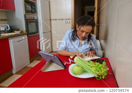 Woman writing notes in kitchen with tablet and healthy snacks Woman writing notes in kitchen with tablet and healthy snacks 120739574