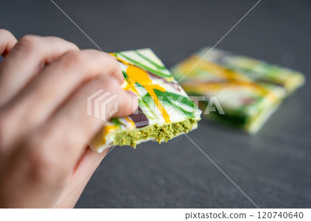 Piece of trend Dubai white chocolate with pistachio paste and kataifi dough in woman hand. Confectionery handmade sweets at home in the kitchen. Piece of trend Dubai white chocolate with pistachio paste and kataifi dough in woman hand. Confectionery handmade sweets at home in the kitchen. 120740640