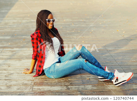Fashion young african woman sitting in a city park, wearing a red checkered shirt and sunglasses Fashion young african woman sitting in a city park, wearing a red checkered shirt and sunglasses 120740744