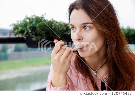 Woman sits savoring her meal with spoon she has long hair is dressed warmly green plants are visible in creating cozy atmosphere. 120745452