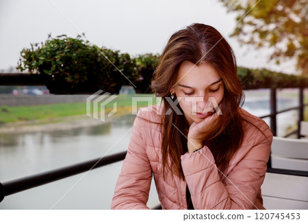 Young woman in light pink jacket rests her chin on her hand, deep in thought while sitting by river atmosphere is calm, with cloudy sky creating reflective mood. Young woman in light pink jacket rests her chin on her hand, deep in thought while sitting by river atmosphere is calm, with cloudy sky creating reflective mood. 120745453