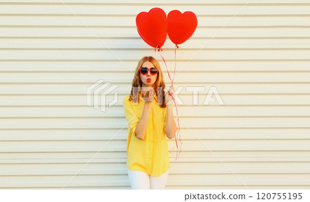 Beautiful young woman with red heart balloon blowing kiss in yellow shirt on white wall background 120755195