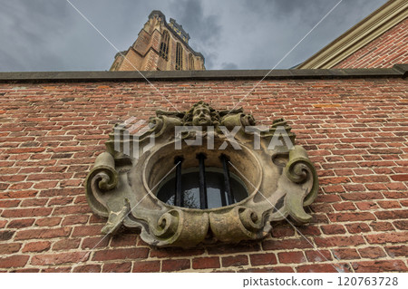 An ornate stone window with decorative carvings on a brick wall, viewed from below, with a towering church or historical structure rising above under a dramatic cloudy sky. 120763728