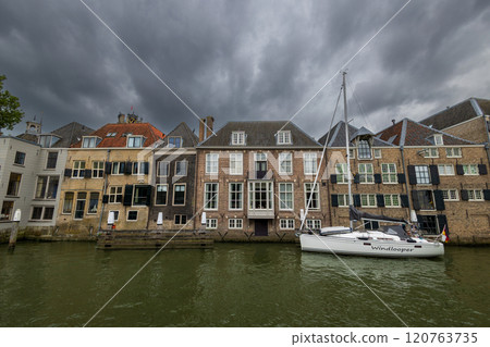 Serene harbor with moored boats under a dramatic cloudy sky. Quaint historic buildings and trees line the waterfront, reflecting in the calm green water, creating peaceful scene. Dordrecht landscape. 120763735