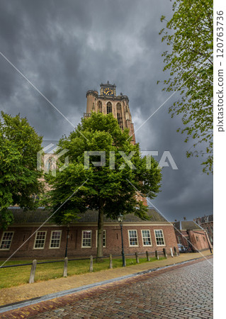 Historic European town with a clock tower under a dramatic cloudy sky. Cobblestone streets lead to a marina filled with boats. Trees and old architecture frame the peaceful scene. Dordrecht landscape Historic European town with a clock tower under a dramatic cloudy sky. Cobblestone streets lead to a marina filled with boats. Trees and old architecture frame the peaceful scene. Dordrecht landscape 120763736