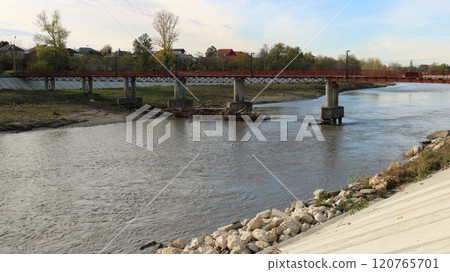 a small red iron bridge over a river with a calm flow of water leading from the bank with a concrete slope and a stone embankment to a residential rural area with trees a small red iron bridge over a river with a calm flow of water leading from the bank with a concrete slope and a stone embankment to a residential rural area with trees 120765701