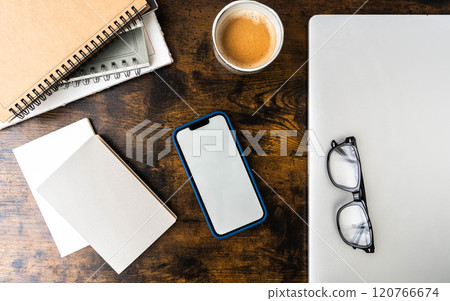 Workspace with smartphone in blue case, spiral notebooks, blank paper, glasses, coffee cup, and laptop on wooden desk 120766674