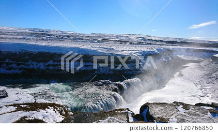 Gullfoss, Iceland - 3.24.2018: A breathtaking view of a roaring waterfall cutting through a pristine snowy landscape and rugged terrain towards icy water under a clear blue sky with sunlight Gullfoss, Iceland - 3.24.2018: A breathtaking view of a roaring waterfall cutting through a pristine snowy landscape and rugged terrain towards icy water under a clear blue sky with sunlight 120766850