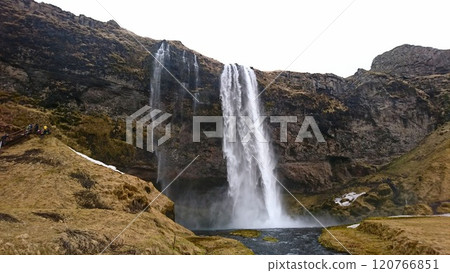 Seljalandsfoss, Iceland - 3.26.2018: Water crashing down from the top of the hill under a cloudy sky with tourists walking on a hiking trail on the cliff around the waterfall before the pandemic 120766851