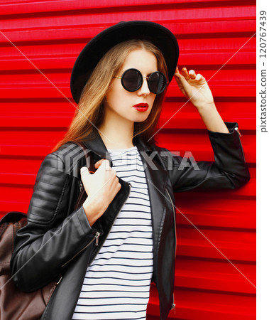 Fashion portrait young woman in black rock style hat, jacket stands with backpack on red background 120767439