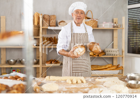 Portrait of elderly baker with bread and baguettes in hands in kitchen 120770914