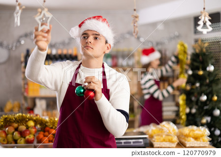 Young guy seller decorates grocery store for christmas Young guy seller decorates grocery store for christmas 120770979