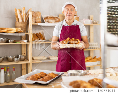 Smiling elderly baker in uniform stands behind counter in small private bakery offering croissants Smiling elderly baker in uniform stands behind counter in small private bakery offering croissants 120771011