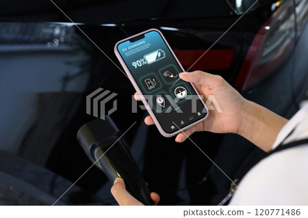 Woman using EV application on smartphone to check charging status while plugged in the EV charging connector 120771486