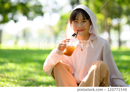Cheerful young woman sitting on the grass in a park and enjoying refreshing drink after exercising 120771548