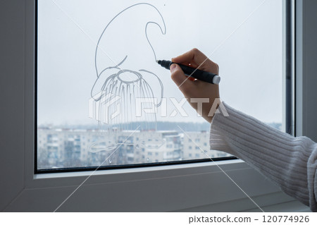 Creative enthusiastic unrecognizable woman in sweater paint window with white marker. Festive gnomes decoration on big clear window. Snowy winter outside 120774926