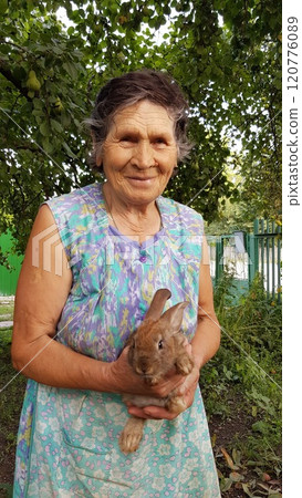 Senior woman holds little rabbit in her elderly hands. Smiling 80s granny in summer garden. Grandmother and brown fur little rabbit 120776089