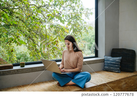 Beautiful woman sitting in front of large window, working on laptop. Beautiful woman sitting in front of large window, working on laptop. 120777678