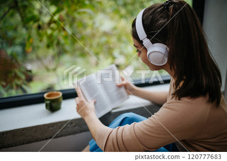 Woman sitting by a large window with headphones on head and book. Woman is enjoying time for herself, reading. 120777683