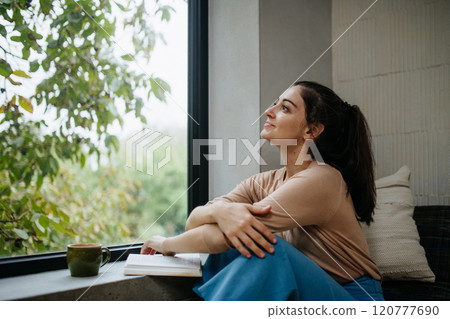 Hygge moment for beautiful woman sitting by a large window with a book. Woman is enjoying time for herself, reading. Hygge moment for beautiful woman sitting by a large window with a book. Woman is enjoying time for herself, reading. 120777690