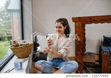 Young businesswoman looking at swatch book, sitting by wooden loom. Upcycled textile, small weaving business and circular economy. Young businesswoman looking at swatch book, sitting by wooden loom. Upcycled textile, small weaving business and circular economy. 120777699