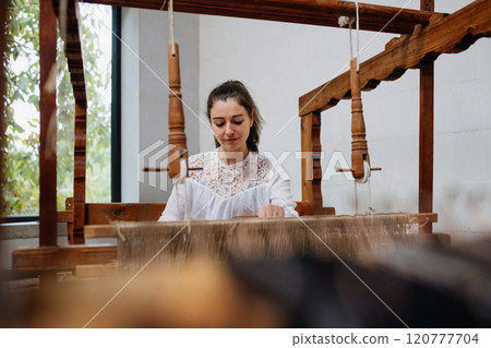 Young businesswoman sitting at a wooden loom, weaving a rug from old fabrics. Upcycling in business and circular economy. Young businesswoman sitting at a wooden loom, weaving a rug from old fabrics. Upcycling in business and circular economy. 120777704