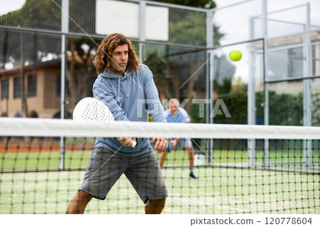 Focused middle-aged Latin man padel player hitting ball with racket on hard court in summer Focused middle-aged Latin man padel player hitting ball with racket on hard court in summer 120778604