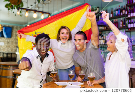 Excited diverse soccer supporters with flag of Spain celebrating victory with pint of beer in the pub 120778605