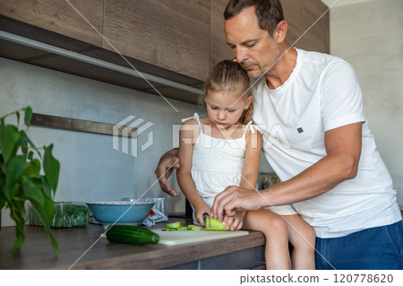 Father patiently teaching a his daughter how to cook in their home kitchen, family with dad and daughter preparing cucumber for green salad with vegetables  120778620
