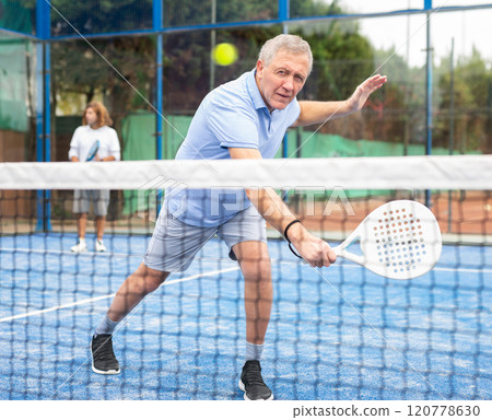 Concentrated elderly woman padel player hitting ball with a racket on hard court 120778630