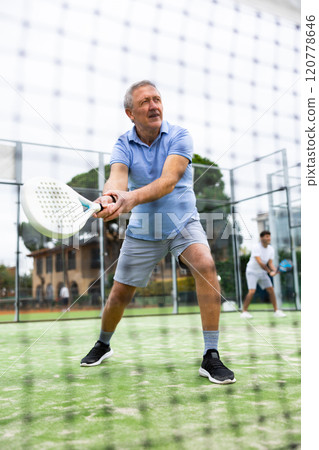 European old man holding playing padel during training in court. View through tennis net 120778646