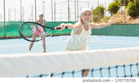 Elderly woman bouncing ball in tennis game on court with racket Elderly woman bouncing ball in tennis game on court with racket 120778708