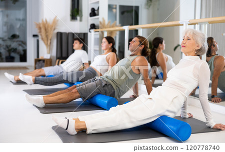 Active senior woman performs exercises with a pilates roller while lying on a mat in the hall of a modern fitness studio. 120778740