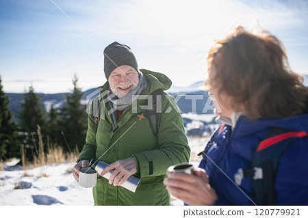 Active seniors resting, drinking warm tea from thermos. Romantic winter hike for an elderly couple in snowy mountains. Active seniors resting, drinking warm tea from thermos. Romantic winter hike for an elderly couple in snowy mountains. 120779921