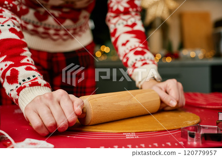 Woman in festive red sweater rolling dough for gingerbread cookies. Process of cooking traditional Christmas sweets for winter holidays at home kitchen Woman in festive red sweater rolling dough for gingerbread cookies. Process of cooking traditional Christmas sweets for winter holidays at home kitchen 120779957