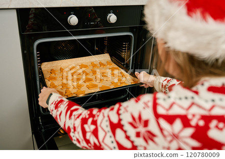 Woman in red festive sweater putting tray of Christmas cookies into oven. Process of cooking traditional gingerbread cookies for winter holidays 120780009
