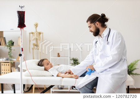 Male doctor provides blood transfusion to young Caucasian girl in medical setting. Child lying on medical bed receiving treatment. Professional healthcare environment, focus on care and comfort. 120782720