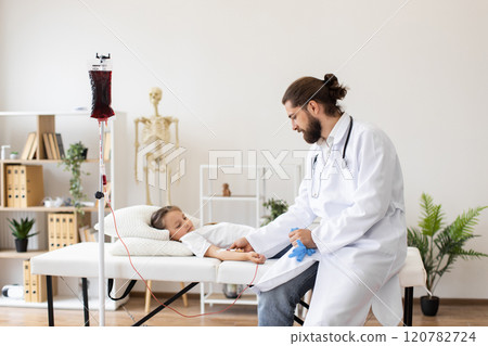 Male doctor gives blood transfusion to young girl at clinic. Scene captures caring medical treatment, wearing lab coat and stethoscope. Plants and medical equipment in background add clinical feel. 120782724