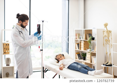 Male doctor provides blood transfusion to young girl of mixed race in clinical setting. Girl lies on examination table, looking calm. Room features medical equipment and skeleton model. 120782739