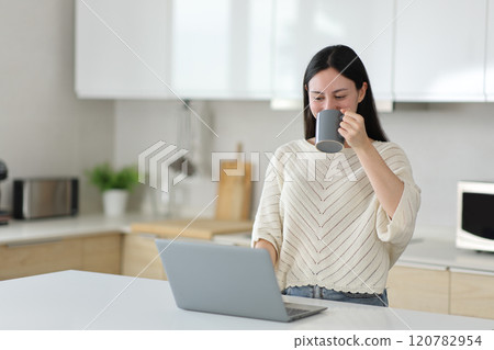 Asian woman drinking and checking laptop in the kitchen 120782954