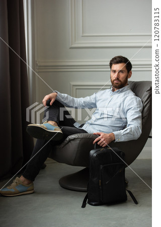 A man is comfortably sitting in a chair, resting with a leather backpack near him A man is comfortably sitting in a chair, resting with a leather backpack near him 120783115