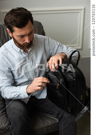 A man is comfortably sitting in a chair, resting with a leather backpack near him 120783116