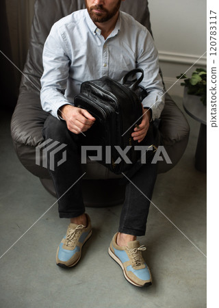 A man is comfortably sitting in a chair, resting with a leather backpack near him 120783117