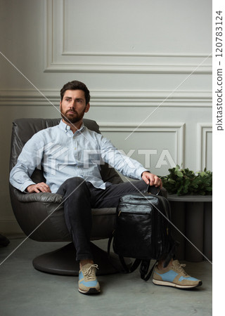 A man is comfortably sitting in a chair, resting with a leather backpack near him 120783124