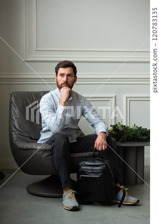 A man is comfortably sitting in a chair, resting with a leather backpack near him A man is comfortably sitting in a chair, resting with a leather backpack near him 120783135