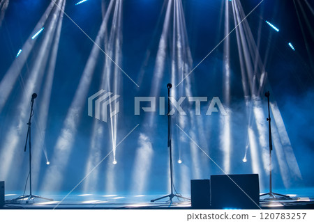 Blurred rays of light from stage spotlights on a blue stage background with microphones. The beginning of a concert or show. 120783257