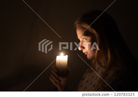 A young woman gazes intently at a lit candle, its warm glow softly illuminating her face against a dark background. The scene evokes a sense of quiet contemplation and intimacy. A young woman gazes intently at a lit candle, its warm glow softly illuminating her face against a dark background. The scene evokes a sense of quiet contemplation and intimacy. 120783325