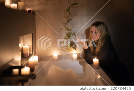 A young woman leans near a bathtub filled with bubbles, holding foam in her hand. The warm glow of candles and soft lighting creates a peaceful and intimate atmosphere A young woman leans near a bathtub filled with bubbles, holding foam in her hand. The warm glow of candles and soft lighting creates a peaceful and intimate atmosphere 120784430