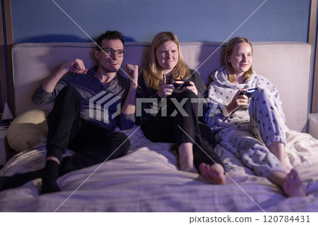 Three friends are sitting on a bed, immersed in playing video games. They are holding controllers, smiling, and enjoying a fun and relaxed gaming session in a cozy atmosphere. 120784431