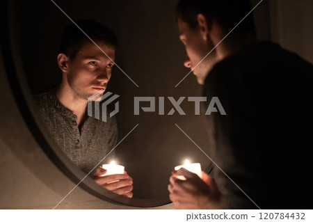 A young man holds a lit candle while gazing at his reflection in a mirror. The soft glow illuminates his thoughtful expression, creating an intimate and introspective atmosphere A young man holds a lit candle while gazing at his reflection in a mirror. The soft glow illuminates his thoughtful expression, creating an intimate and introspective atmosphere 120784432